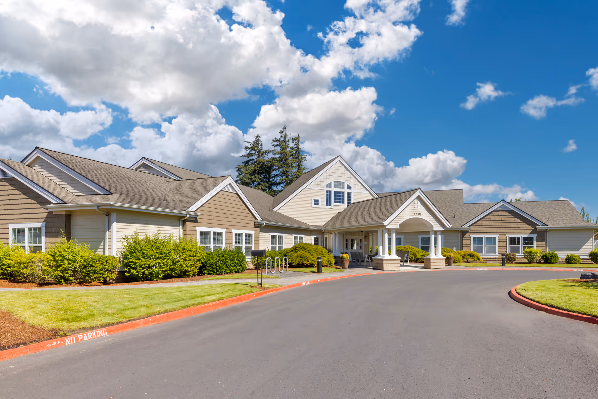 Exterior view of a single-story senior living facility building with beige siding and brown shingles, surrounded by green bushes and a well-maintained lawn under a partly cloudy blue sky. The entrance features a covered porch with white columns and seating.