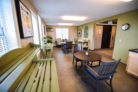 A bright and cozy common area with green walls and large windows letting in natural light. The room features a green bench along the left wall, a round table with chairs in the center, and a seating area with armchairs and a small table near the back. There are framed pictures on the walls, a clock, and a small wooden table near an elevator door on the right side.