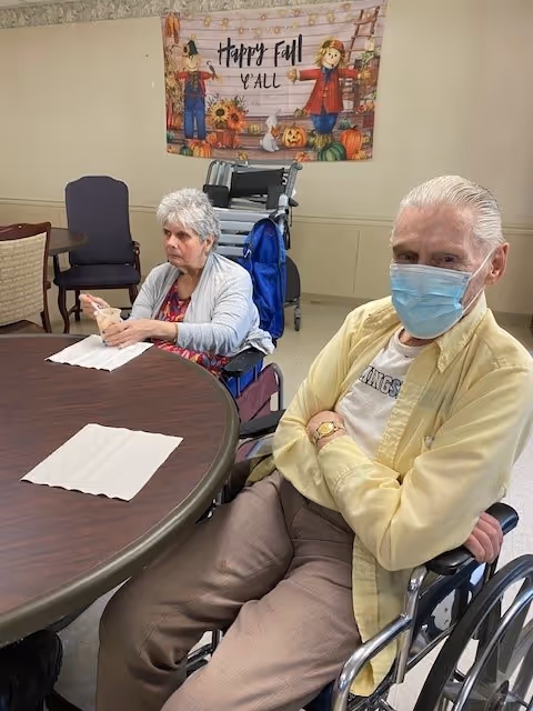 Two elderly individuals sitting in wheelchairs at a round table in a room. One person is wearing a yellow shirt and a blue face mask, while the other person is wearing a gray cardigan. Behind them is a festive fall-themed banner on the wall that reads 'Happy Fall Y'all' with scarecrows, pumpkins, and autumn decorations.