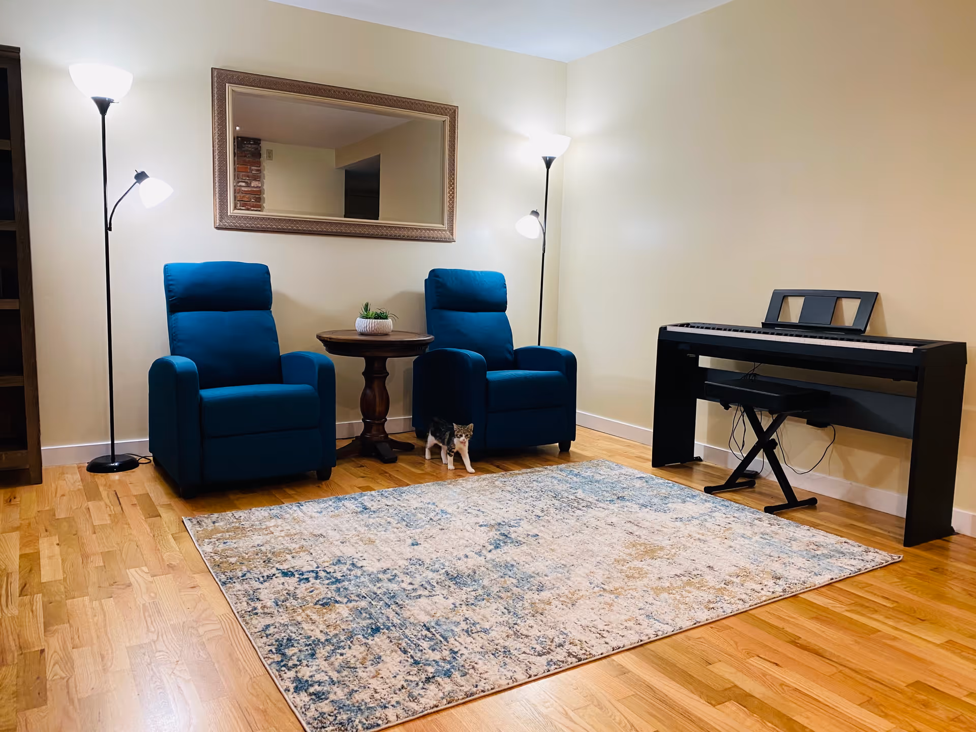A cozy living room with two blue armchairs separated by a small round wooden table with a plant on it. There are two floor lamps behind each chair providing warm light. A large rectangular mirror hangs on the wall above the chairs. On the right side, there is a black digital piano with a matching bench. A patterned area rug covers part of the wooden floor, and a small cat is standing on the floor near the chairs.