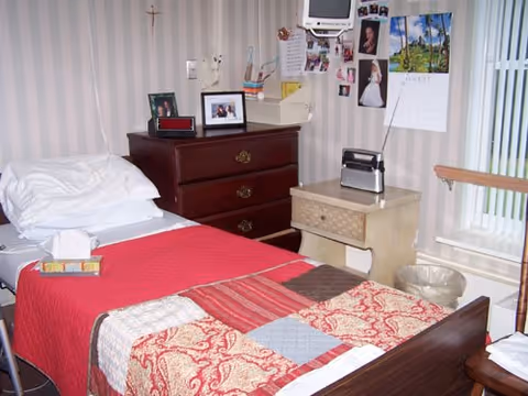 Small bedroom with a single bed covered by a red patterned quilt, a wooden dresser, nightstand, and personal photos on the wall.