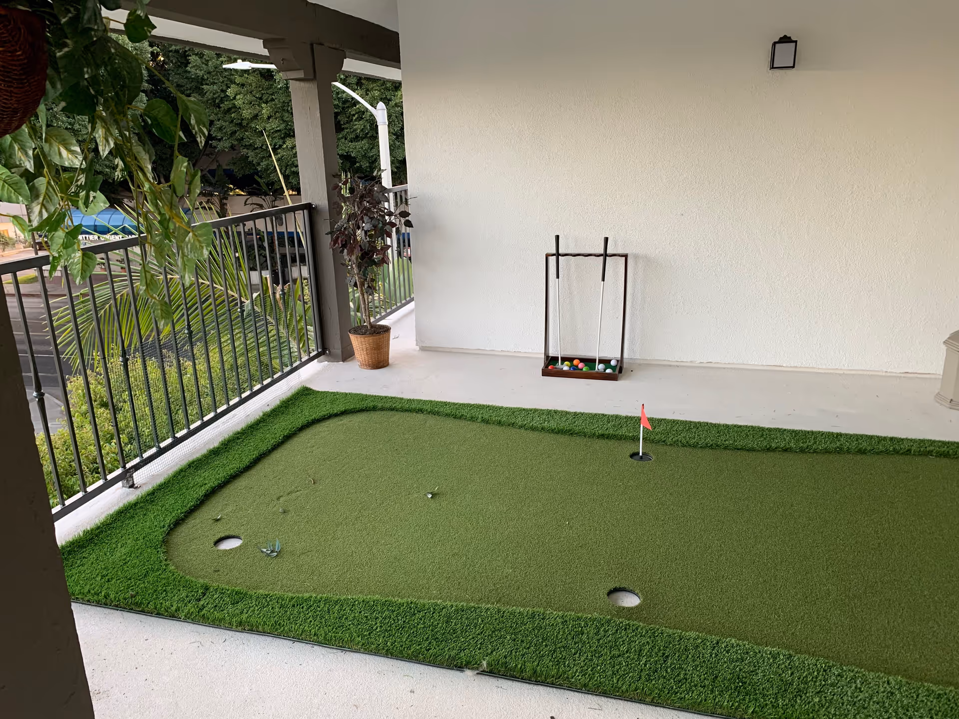 Covered outdoor balcony area with a small putting green mat, a set of croquet mallets and balls in a wooden holder, potted plants, and a metal railing overlooking greenery and a street.
