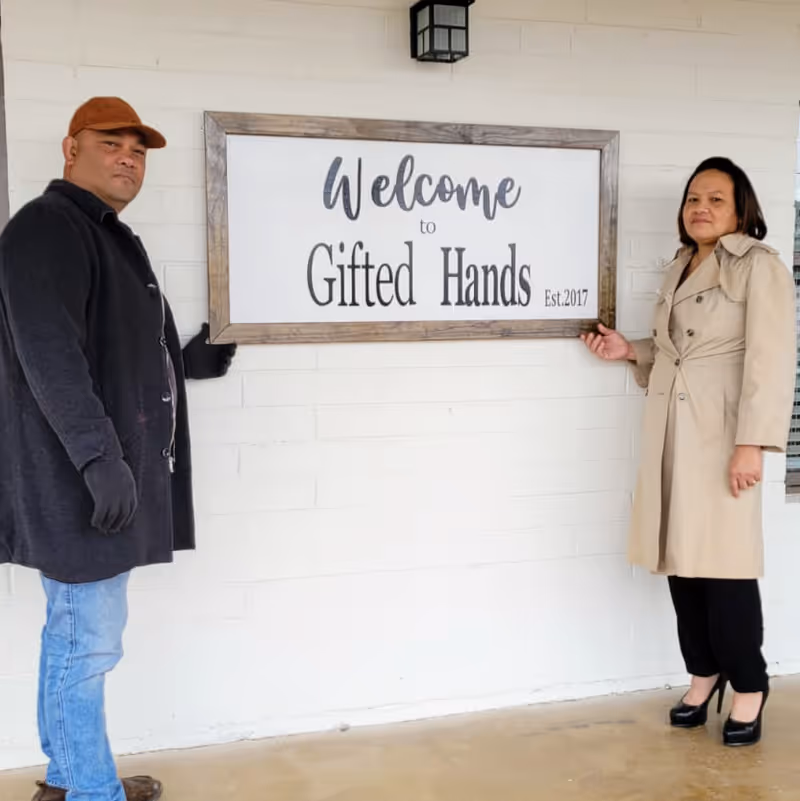 Two people stand on either side of a framed 'Welcome to Gifted Hands' sign mounted on a white brick exterior wall.