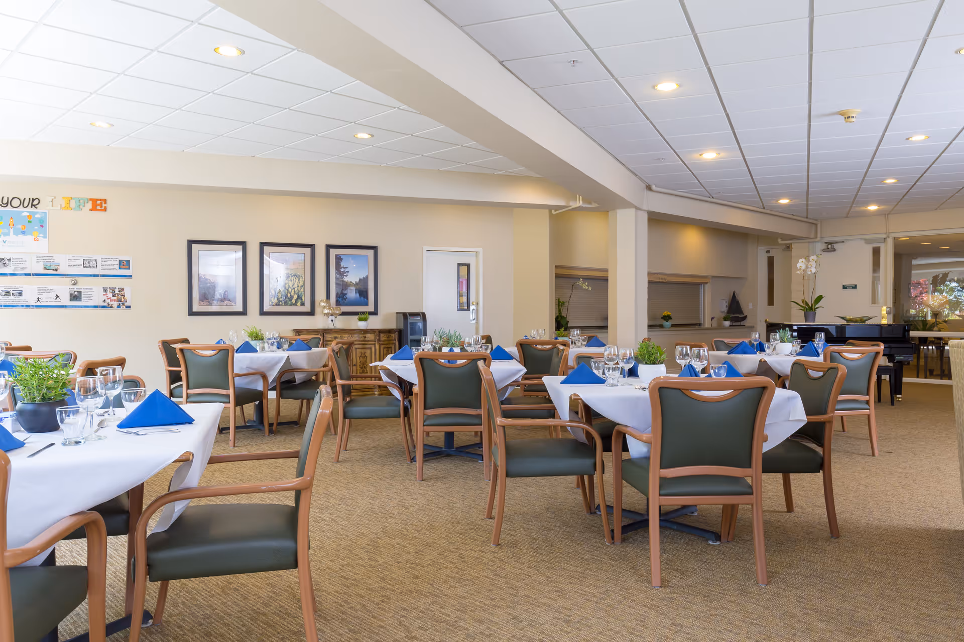 A well-lit dining room with several tables covered in white tablecloths, each set with blue napkins, glassware, and small potted plants. The room has beige walls adorned with framed pictures and informational posters. The floor is carpeted, and there are green cushioned chairs with wooden frames around the tables. In the background, there is a piano and a door leading to another area.