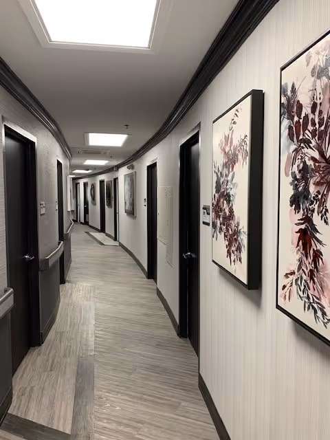 A curved hallway in a senior living facility with light-colored walls and wood-patterned flooring. Several closed dark wooden doors line both sides of the hallway. The walls are decorated with framed floral artwork. Ceiling lights illuminate the corridor.