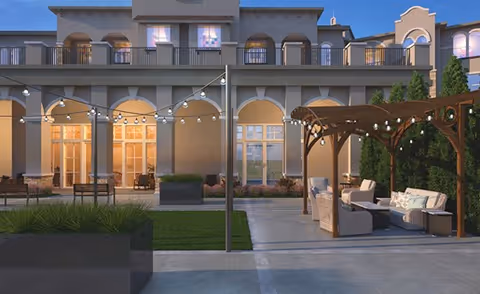 Outdoor patio area of a senior living facility with string lights hanging above, a pergola with cushioned seating, and a building facade with large windows and balconies in the background during dusk.