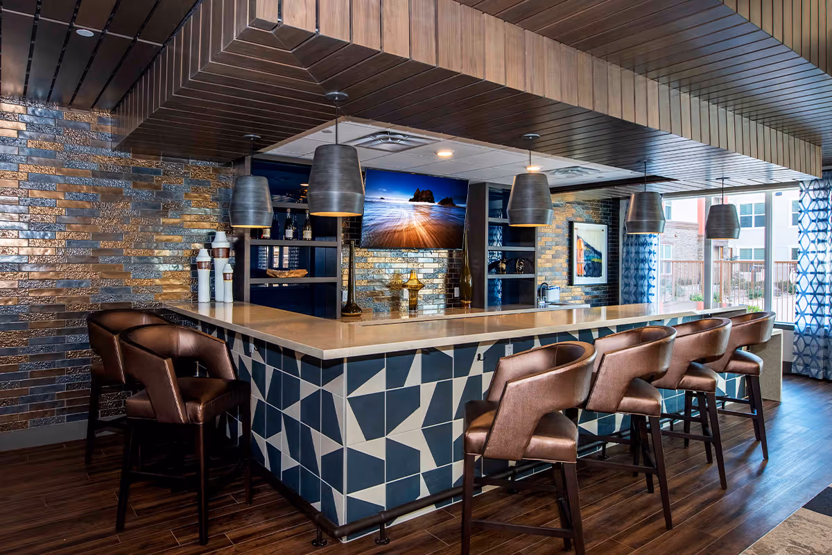 Modern bar area with a geometric patterned counter, six brown leather bar stools, pendant lights hanging from a wooden ceiling, a metallic tiled wall, and a flat-screen TV mounted on the wall. Large windows with blue patterned curtains allow natural light into the space.