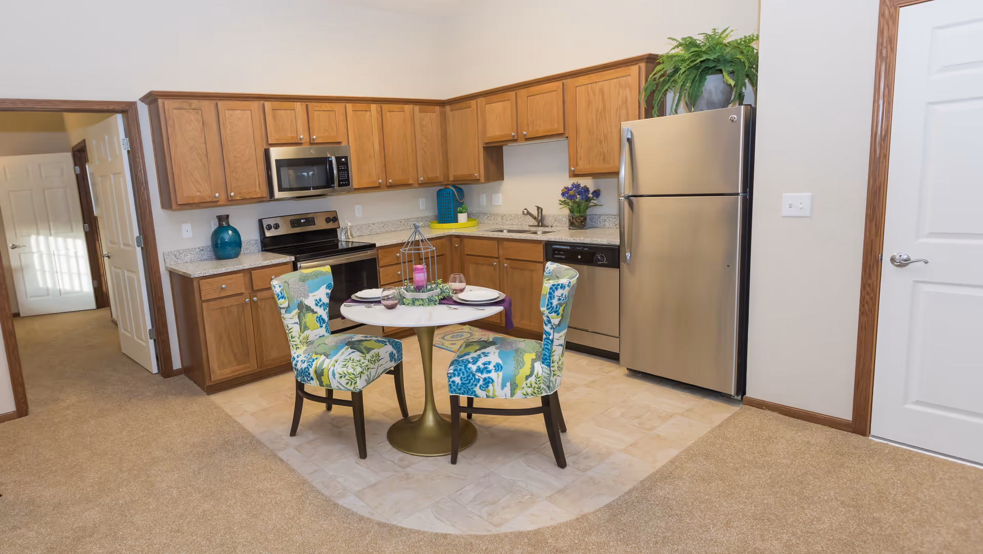 A kitchen area in a retirement facility featuring wooden cabinets, a stainless steel refrigerator, stove, microwave, and dishwasher. In the center, there is a small round dining table with two colorful patterned chairs. The floor transitions from carpet to tile in the kitchen area, and there are decorative items like a plant on top of the refrigerator and a vase with flowers on the counter.
