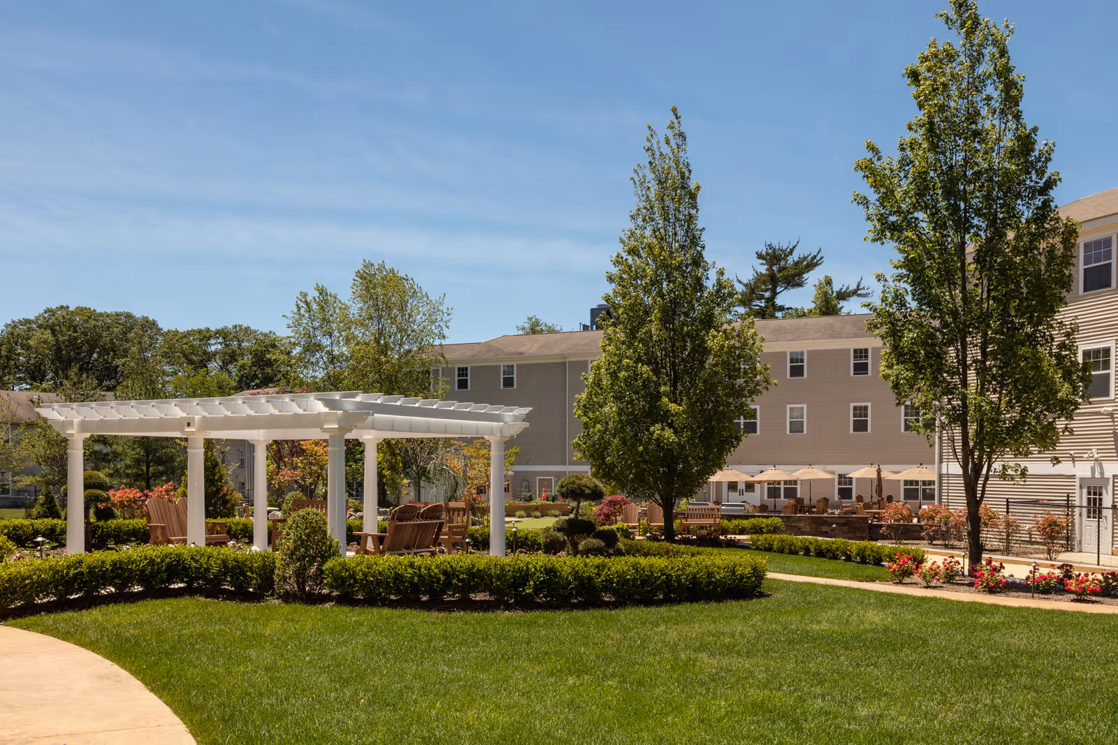 Outdoor garden area at an assisted living facility with a white pergola, wooden chairs, green grass, trees, and a beige multi-story building in the background under a clear blue sky.