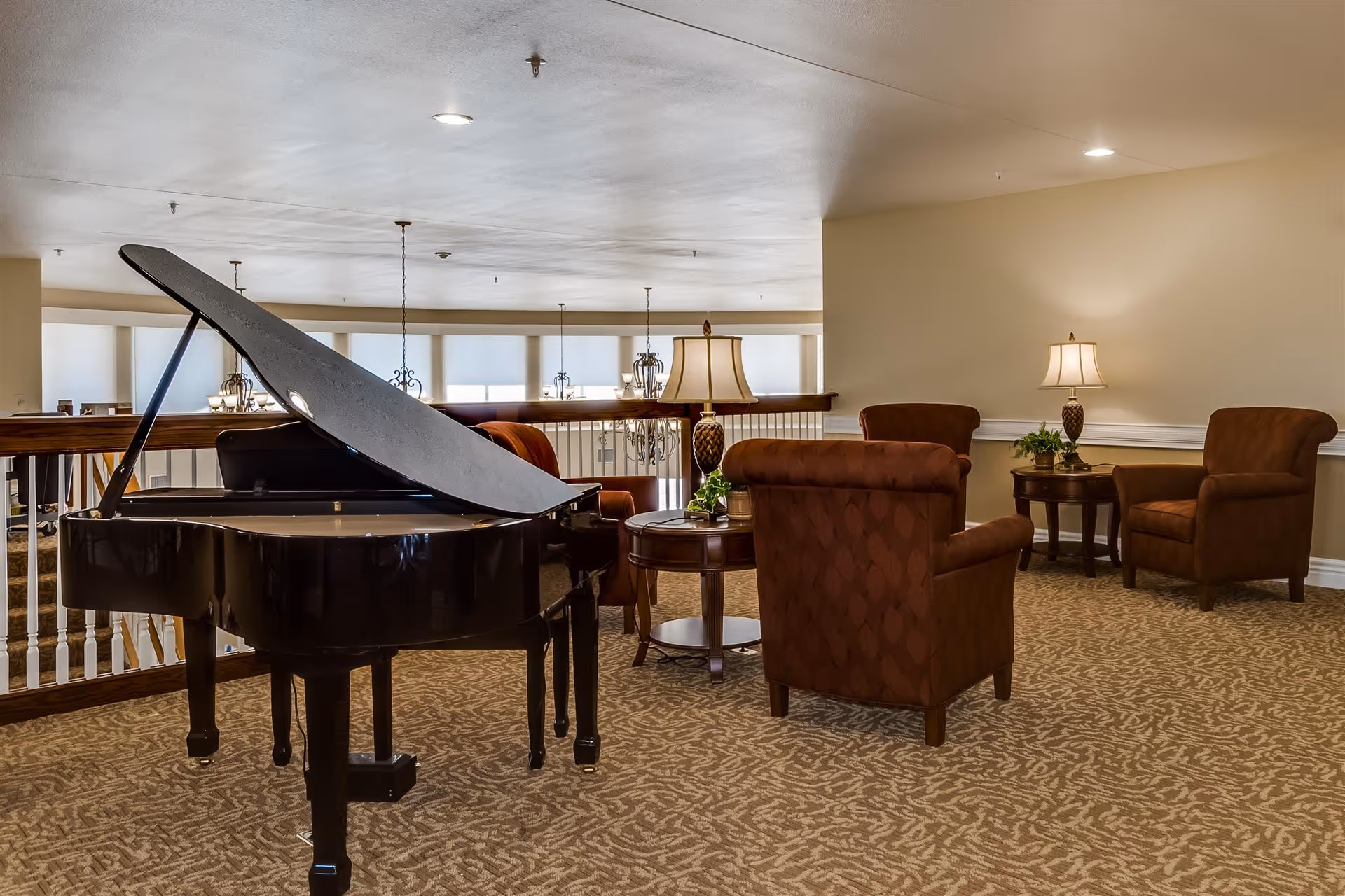 A cozy interior lounge area featuring a black grand piano, two round wooden tables with table lamps, and several upholstered armchairs in a warm brown color. The space has carpeted flooring with a patterned design and a railing overlooking a lower level with large windows and hanging light fixtures.