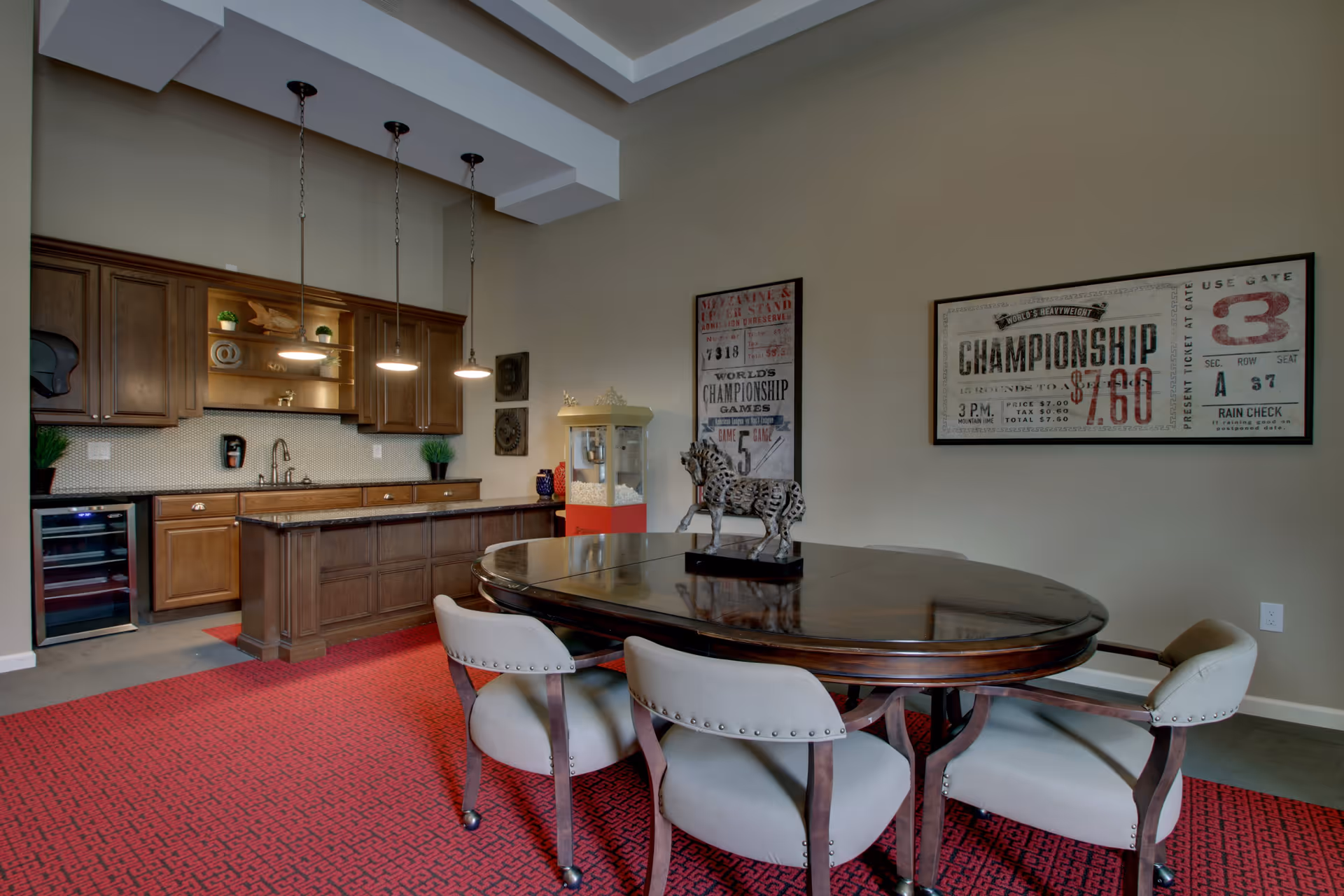 Interior view of a senior living facility room featuring a dark wooden oval table with four upholstered chairs on a red patterned carpet. In the background, there is a kitchenette with wooden cabinets, a sink, a small wine fridge, and three pendant lights hanging from the ceiling. Two framed vintage-style championship posters and a popcorn machine are visible on the beige walls.
