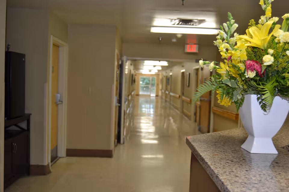 A long interior hallway of a care facility with handrails and a vase of yellow flowers on a front counter in the foreground.