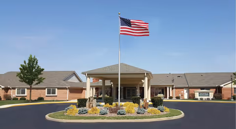 Front entrance of a single-story brick senior living facility with a circular driveway, landscaped island and an American flag on a flagpole.