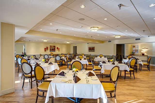 A spacious dining room with multiple tables covered in white tablecloths, each set with plates, silverware, glasses, and folded brown napkins. The chairs have yellow cushioned seats and backs with dark wooden frames. The room has wooden flooring, soft yellow walls, ceiling lights, and framed artwork on the walls.