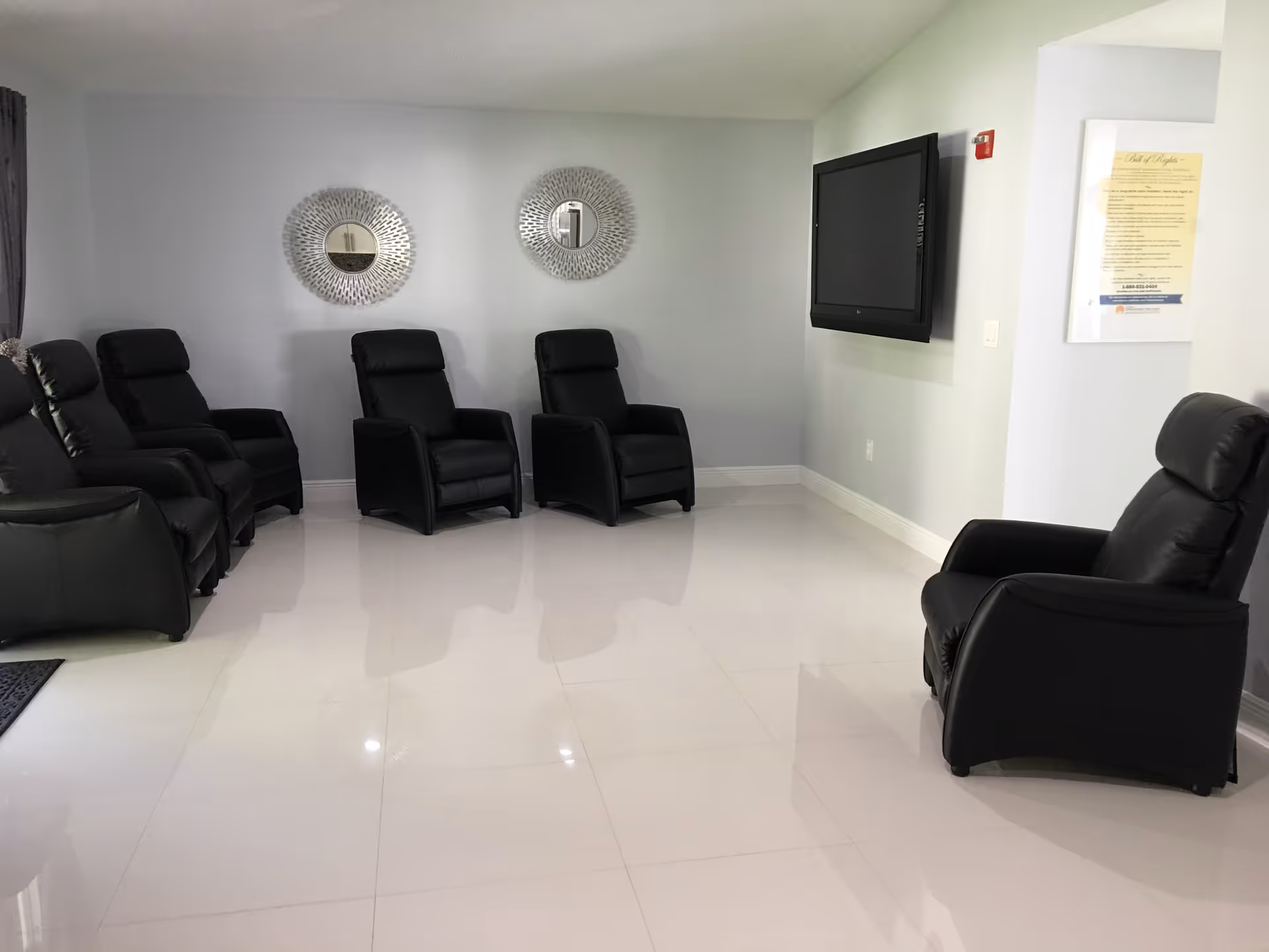 A seating area with black reclining chairs and a television on the wall in a senior living facility.