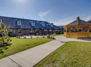 Outdoor view of a senior living facility with a stone building featuring multiple dormer windows, a paved walkway, green grass, small trees, and a wooden gazebo under a clear blue sky.