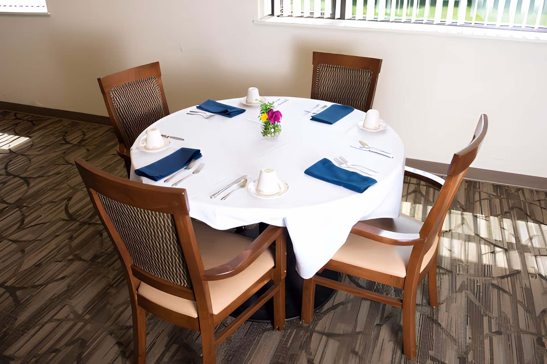 Round dining table set for four with a white tablecloth, blue napkins, and wooden chairs in a dining room.