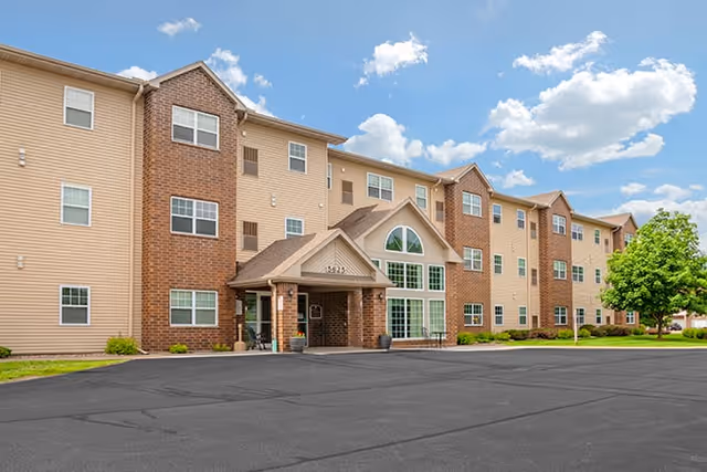 Exterior view of a three-story senior living facility building with beige siding and brick accents, a covered entrance, multiple windows, and a paved parking area in front under a partly cloudy sky.