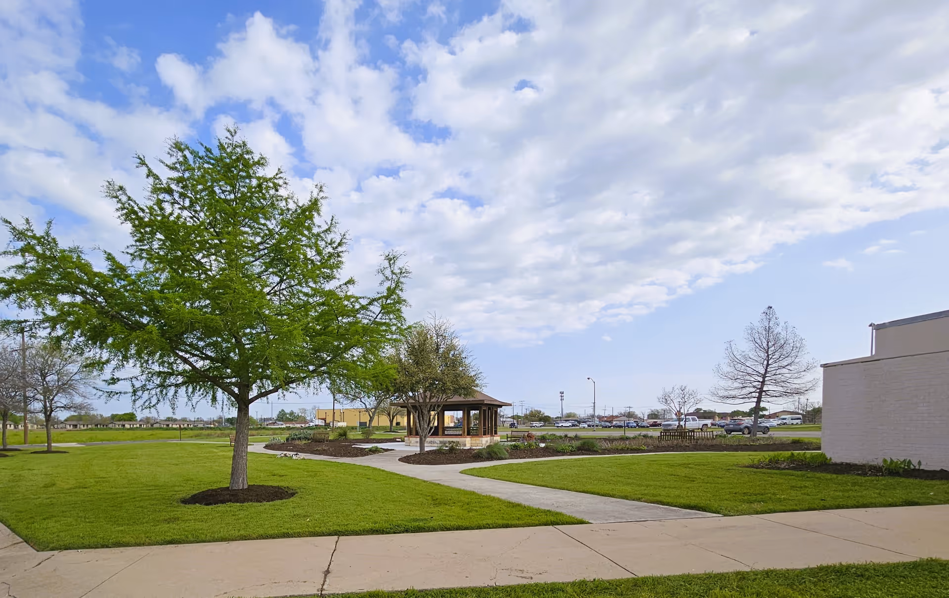 A landscaped lawn with a paved walkway, trees and a small gazebo under a partly cloudy sky.