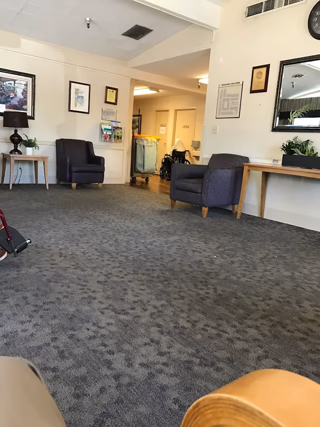 Carpeted common room in a care center with armchairs, side tables, framed artwork, and a hallway holding a wheelchair and a laundry cart.