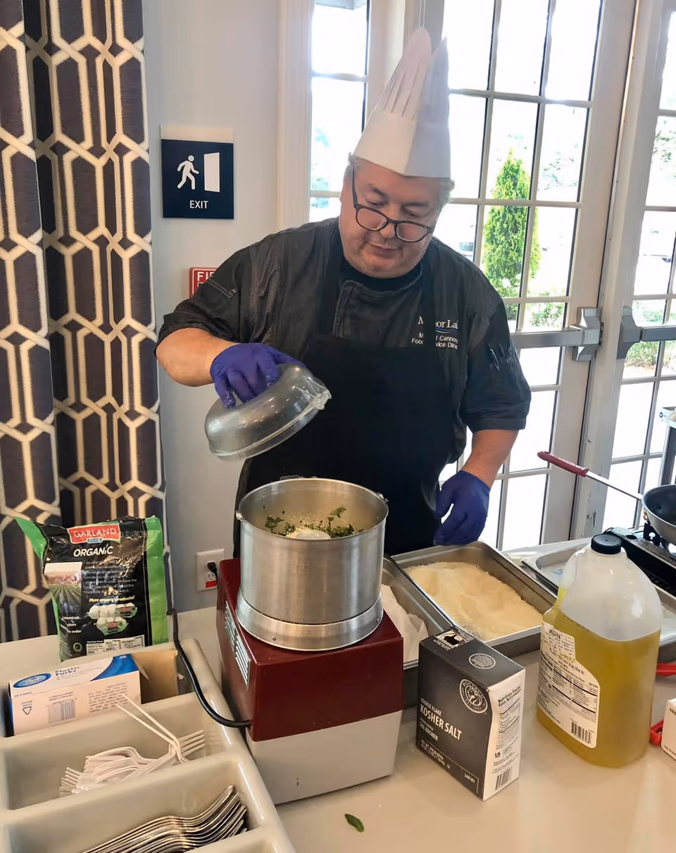 A chef wearing a white paper hat, black apron, and purple gloves is preparing food using a food processor on a table. Various ingredients and kitchen items, including a bag of organic greens, kosher salt, a large container of oil, and trays of food, are visible on the table. The setting appears to be a bright room with large windows and a patterned curtain.