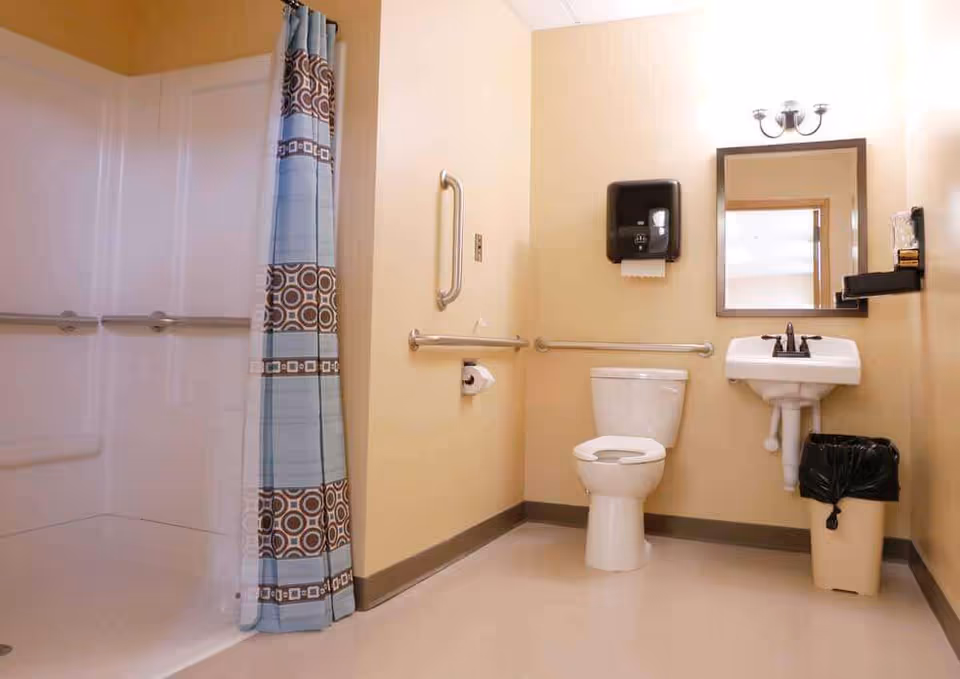 A clean and accessible bathroom with beige walls, featuring a white toilet, a white sink with a mirror above it, a trash can, a paper towel dispenser, and a shower area with a blue and brown patterned shower curtain. Grab bars are installed near the toilet and inside the shower for support.