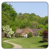 A scenic outdoor view of a pathway curving through a well-maintained grassy area with trees and shrubs, leading to a brick building with a sloped roof. Two people are walking on the path towards the building under a clear blue sky.