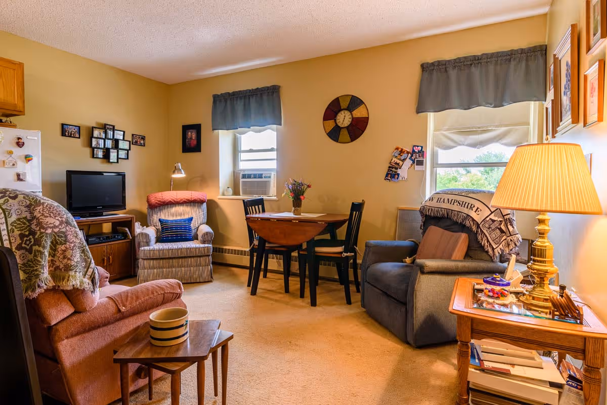 A cozy living room in a senior living facility with two armchairs, a small wooden table with a striped mug, a round dining table with four chairs, a TV on a wooden stand, and a side table with a lamp. The room has beige walls, two windows with blue valances, and various framed pictures and decorations on the walls.