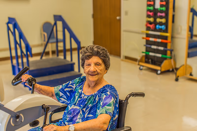 An elderly woman in a wheelchair using a hand cycle exercise machine in a rehabilitation room with exercise equipment and stairs in the background.