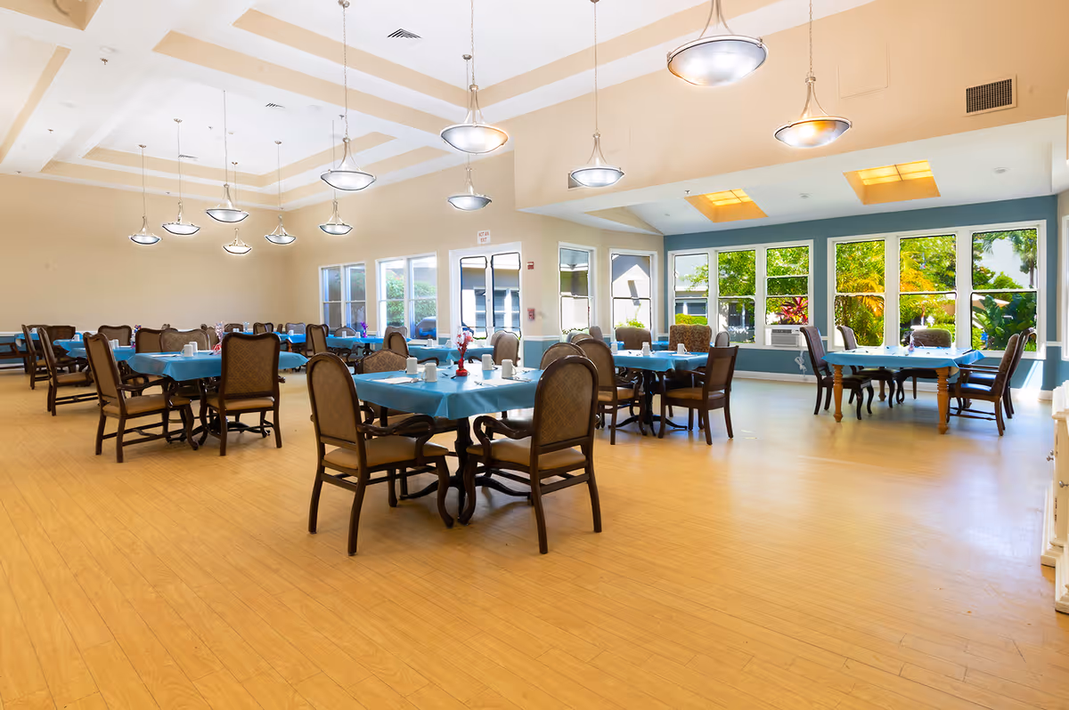 Bright, spacious dining room with multiple tables covered in blue tablecloths and large windows overlooking greenery.