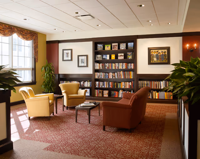 A cozy library or reading room with a large bookshelf filled with books against the wall. The room has two yellow armchairs and one red armchair arranged around a small coffee table with magazines. There are framed pictures on the walls, large windows with patterned valances letting in natural light, and green plants adding a touch of nature. The carpet has a red and beige pattern, and the ceiling has recessed lighting.