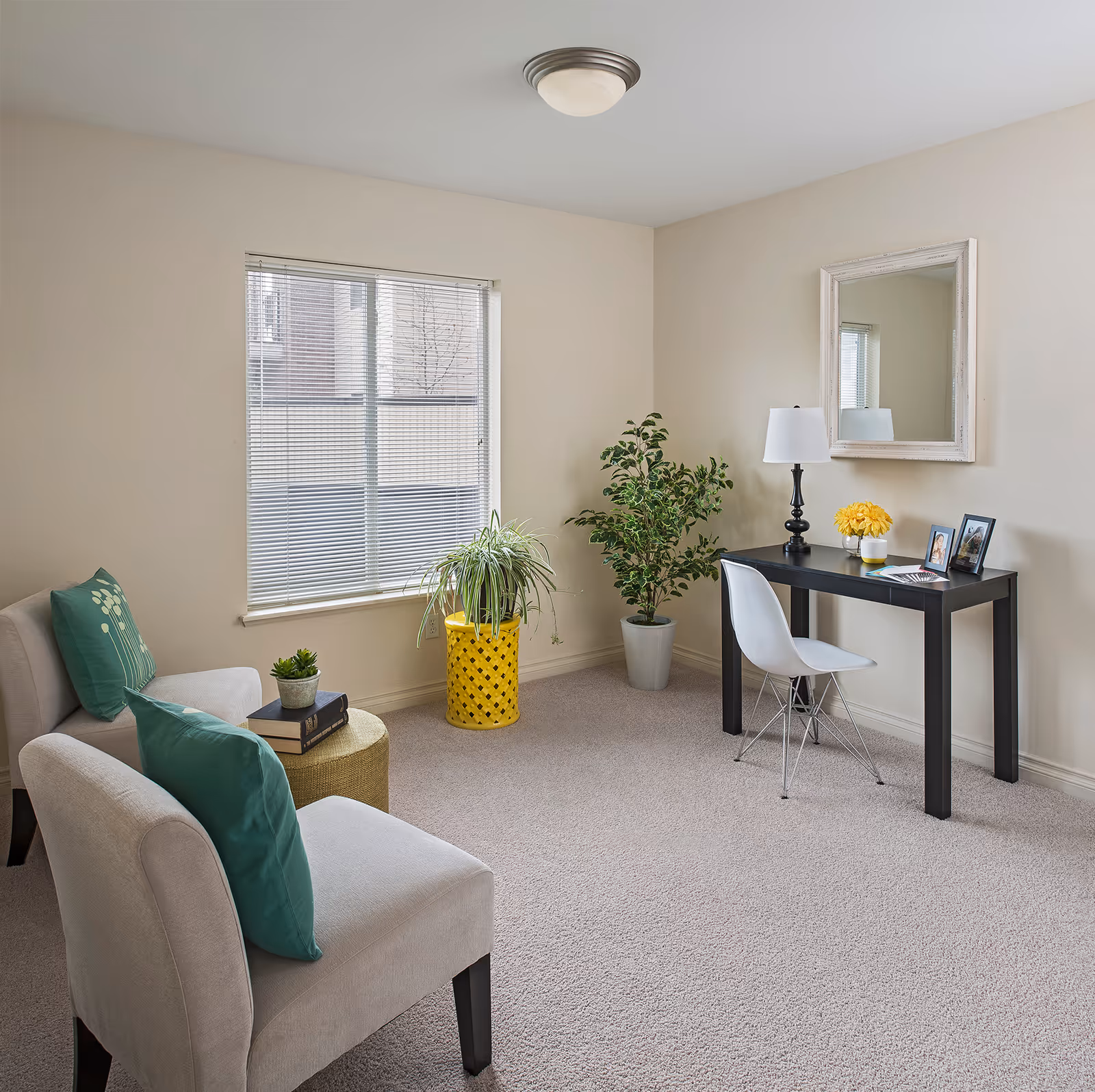 A cozy interior room with two beige chairs each with a green cushion, a small round ottoman with two books and a small plant on it, a window with blinds, two potted plants, and a black desk with a white chair. On the desk are a table lamp, a yellow flower arrangement, and two framed photos. A mirror hangs on the wall above the desk.