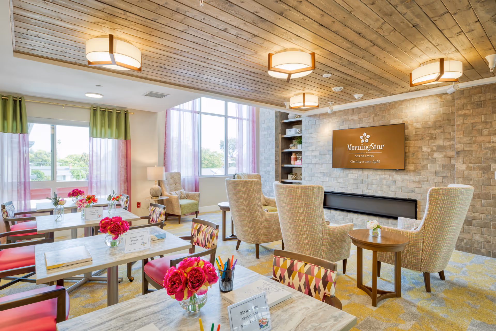 Bright senior living common room with dining tables, colorful chairs, lounge armchairs, and a wall-mounted TV.