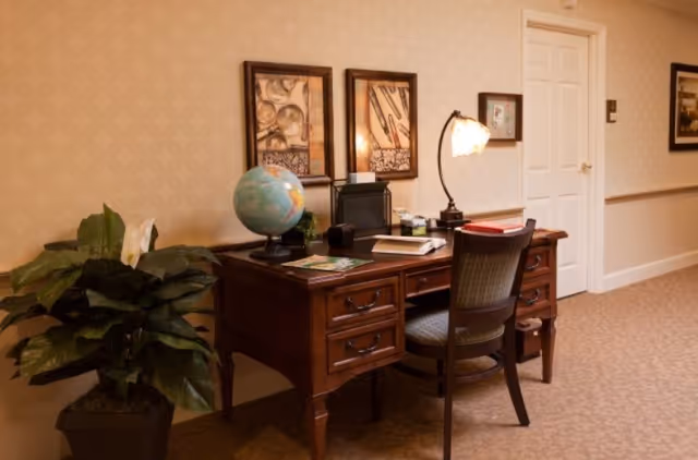 A wooden desk and chair in a carpeted interior hallway, topped with a globe, lamp, books and framed artwork on the wall.