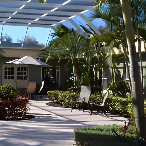 Outdoor patio area with a pathway surrounded by greenery, bushes, and trees. There are benches along the path and a small building with windows and a white umbrella providing shade over a seating area.