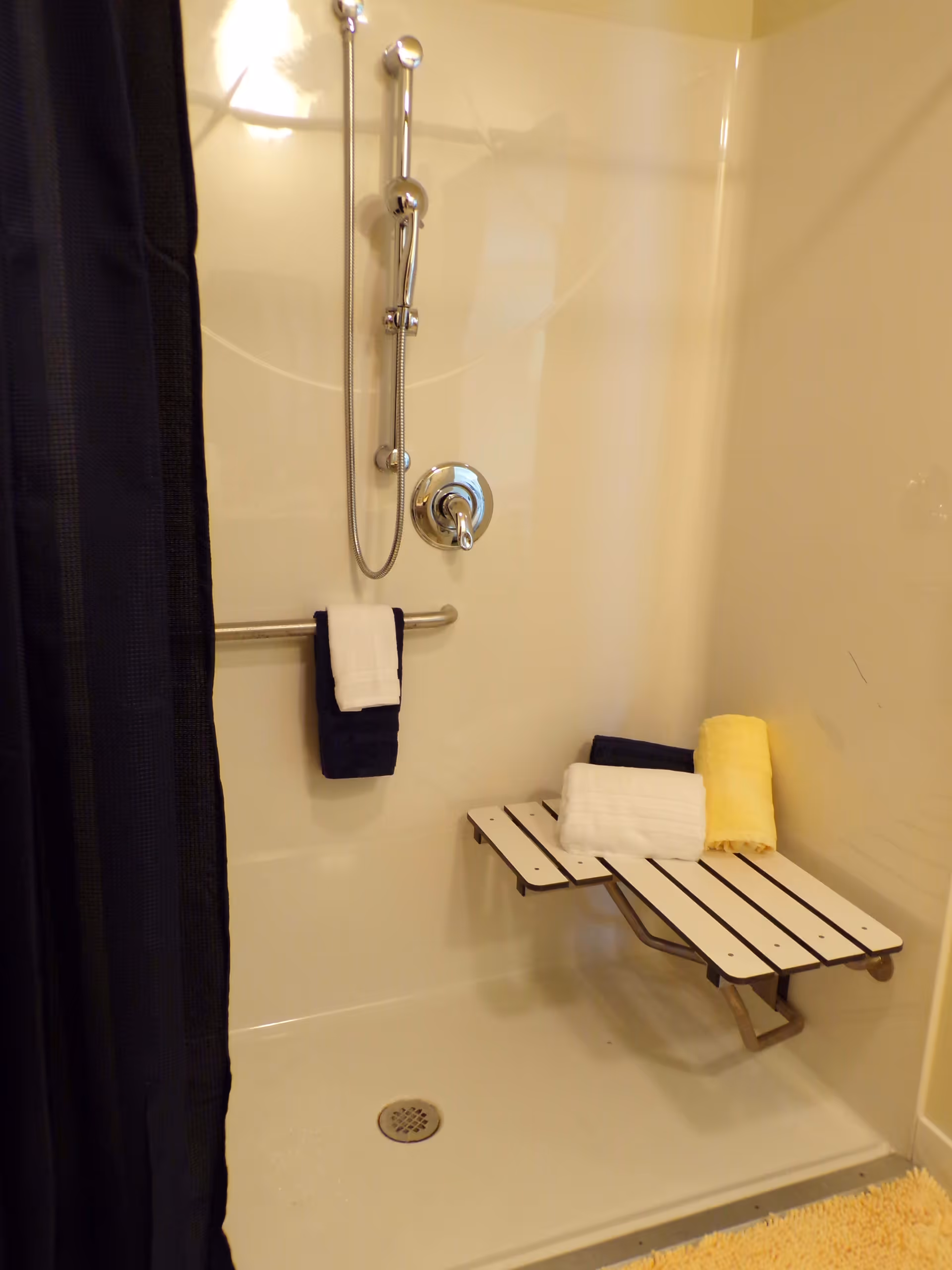 Shower area with a black shower curtain, a handheld showerhead, a metal grab bar with two towels hanging on it, and a fold-down shower seat holding three rolled towels in white, yellow, and dark blue.