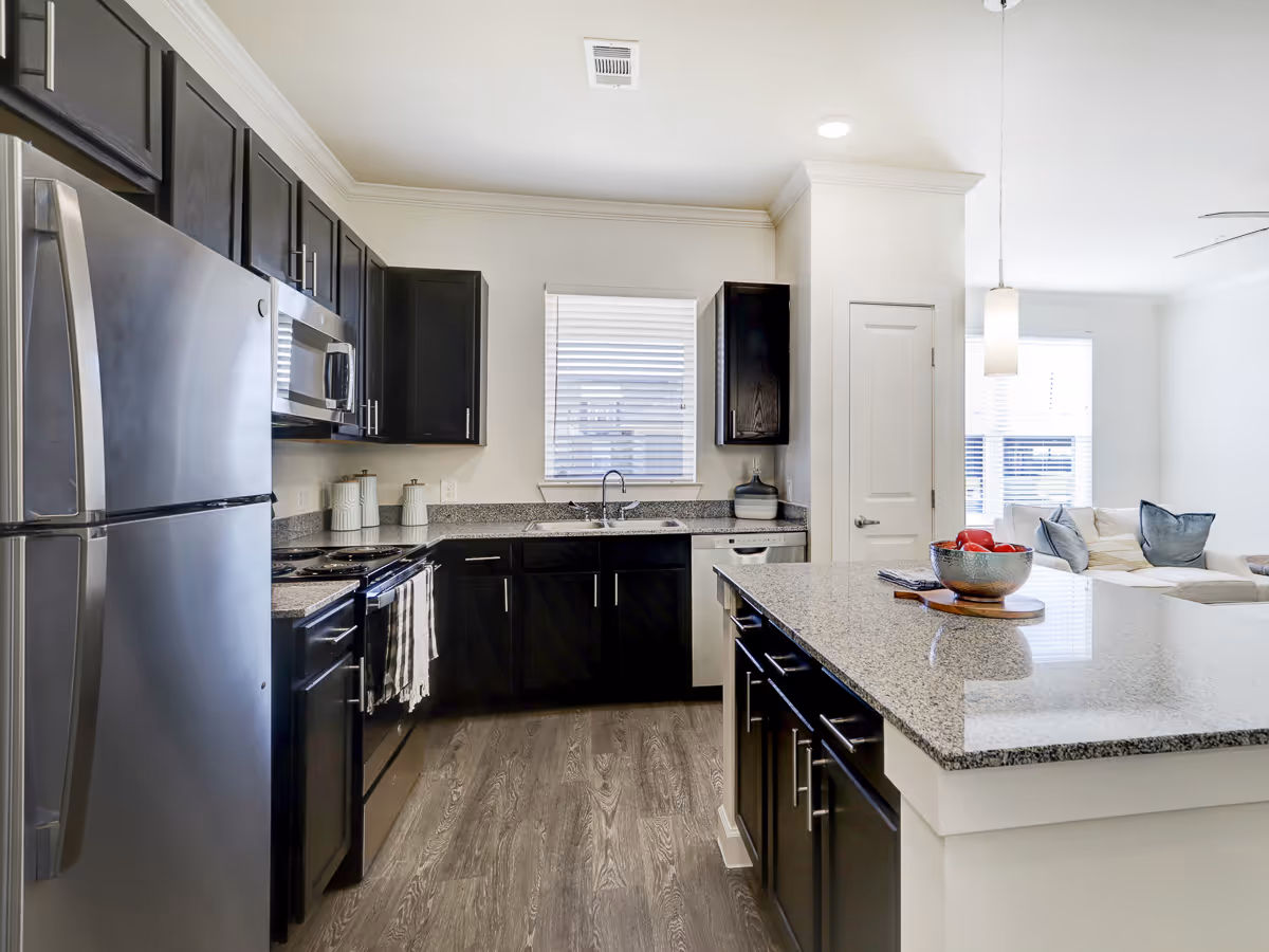Modern kitchen with dark wood cabinets, stainless steel refrigerator, microwave, oven, and dishwasher. Granite countertops with a large island in the center, a bowl of red apples on the island, and a window above the sink letting in natural light. The kitchen opens into a living area with a white couch and pillows.