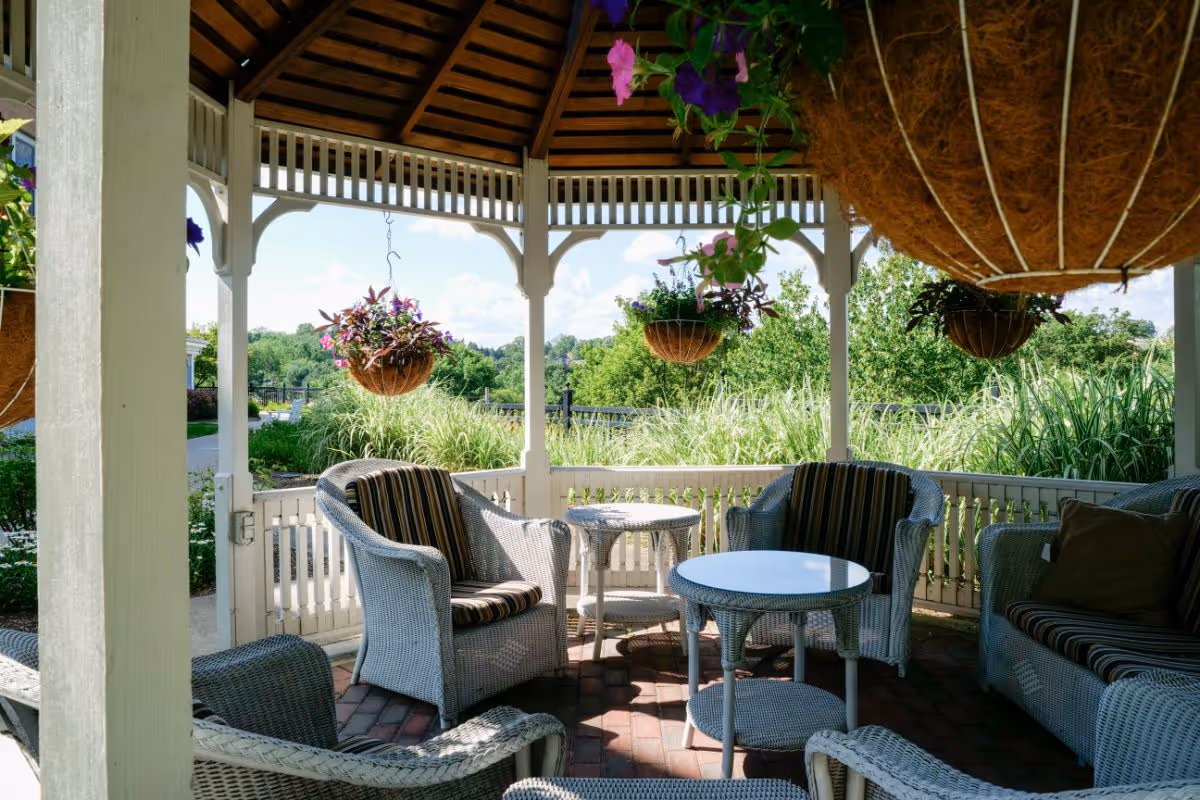 Wicker chairs and small tables arranged inside a wooden gazebo with hanging flower baskets and lush greenery beyond.