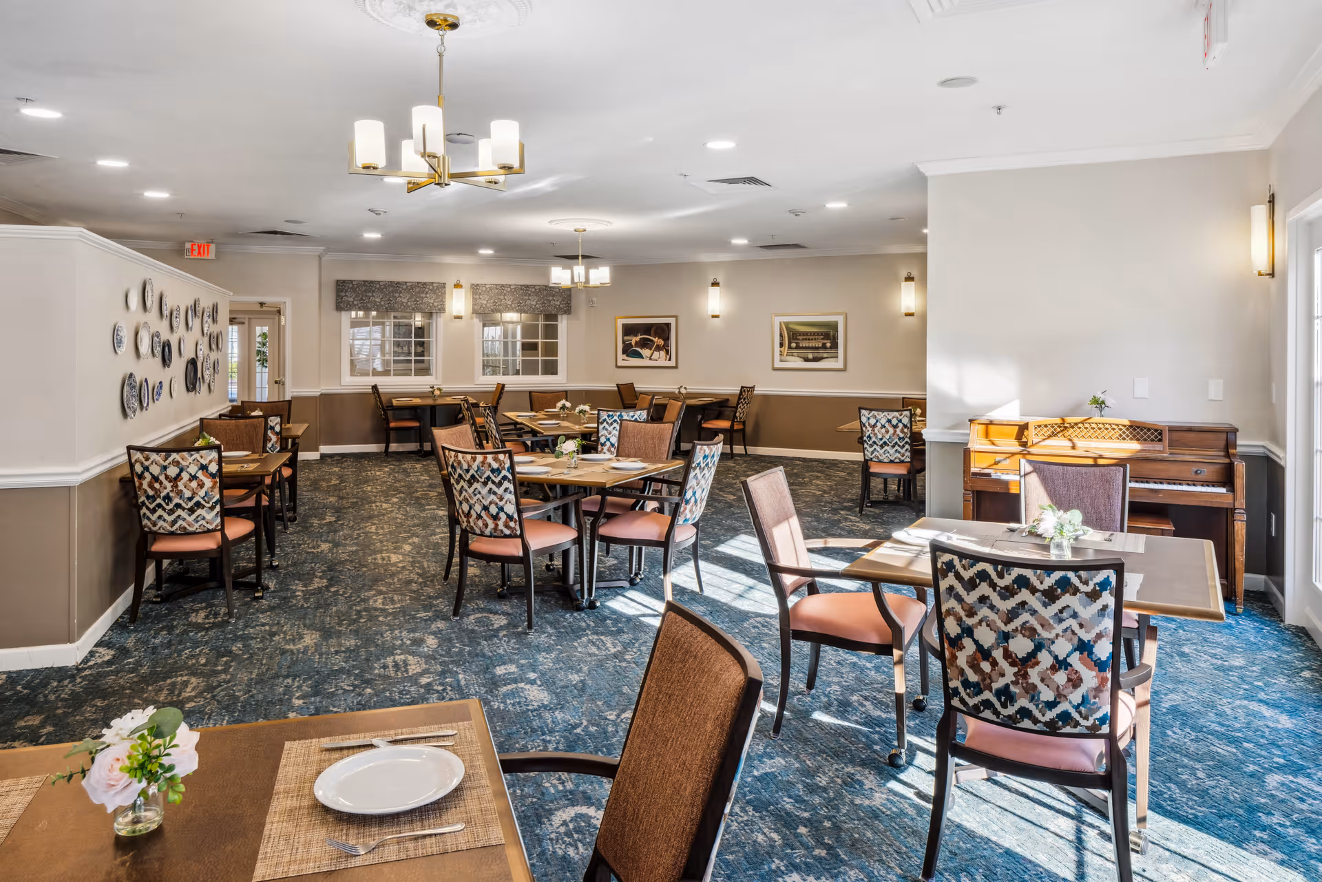 Bright dining room with multiple set tables and patterned chairs, a piano at right, and decorative plates on the wall.