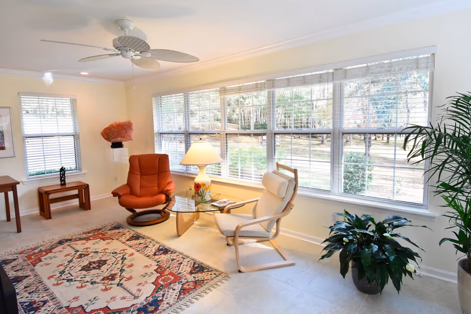 A bright living room with large windows covered by white blinds, allowing natural light to fill the space. The room features a red cushioned swivel chair, a light wooden armchair with white cushions, a glass coffee table with a floral lamp on it, a patterned area rug, two potted plants, and a ceiling fan with leaf-shaped blades.