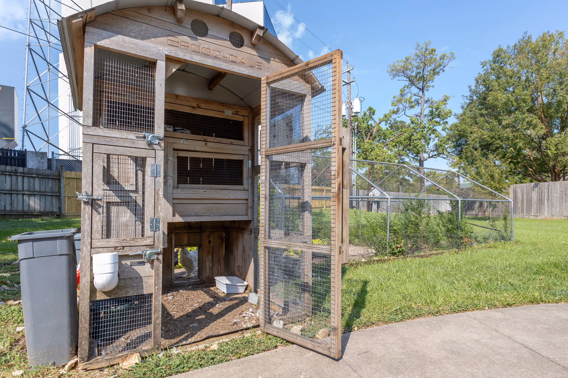 Outdoor wooden chicken coop with wire mesh doors open, situated on a grassy area next to a paved walkway. A white chicken is visible inside the coop. Trees and a fenced garden area are in the background under a clear blue sky.