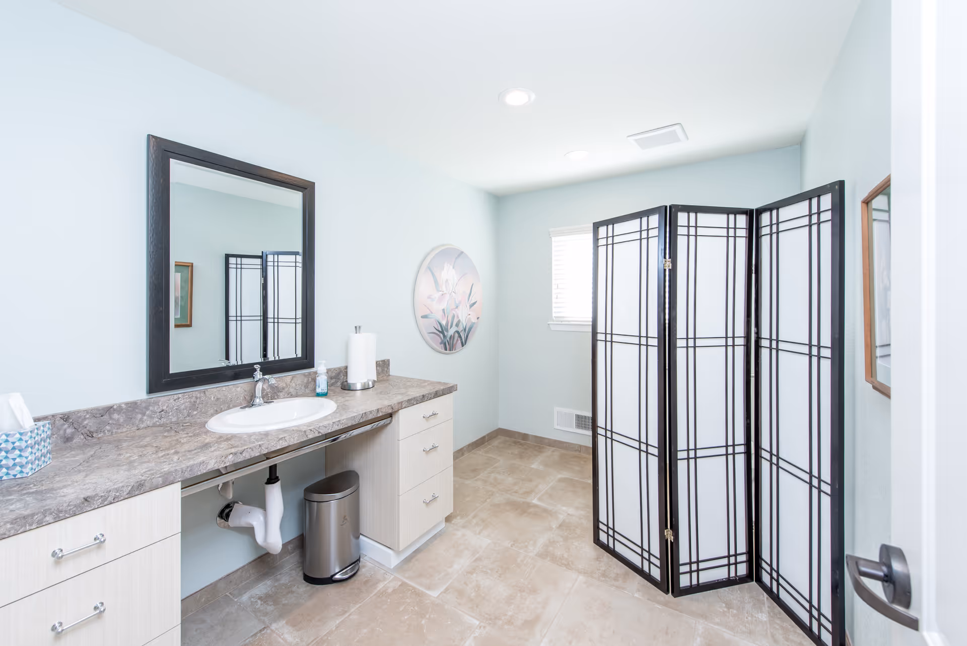 Bright accessible bathroom with a countertop sink and mirror, a folding privacy screen, and tiled floor.