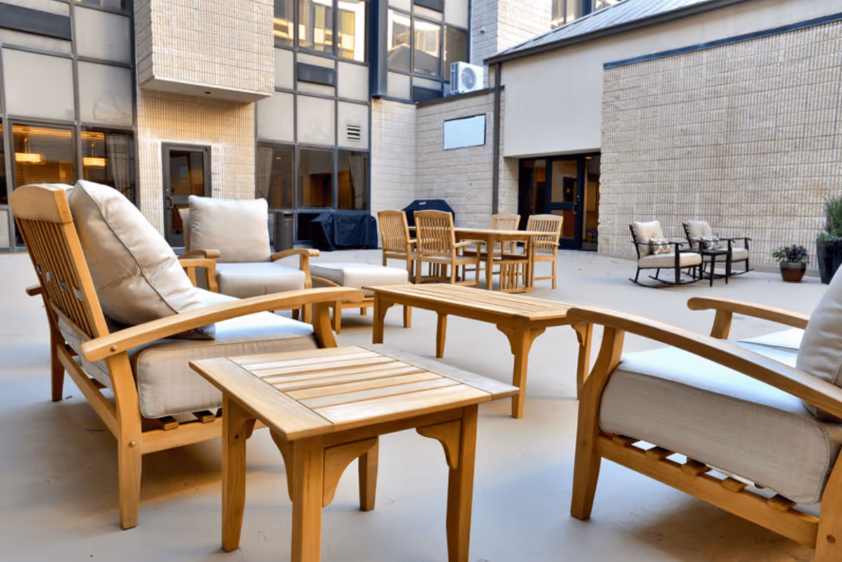 Outdoor patio area with wooden chairs and tables featuring beige cushions, surrounded by a building with large windows and doors.