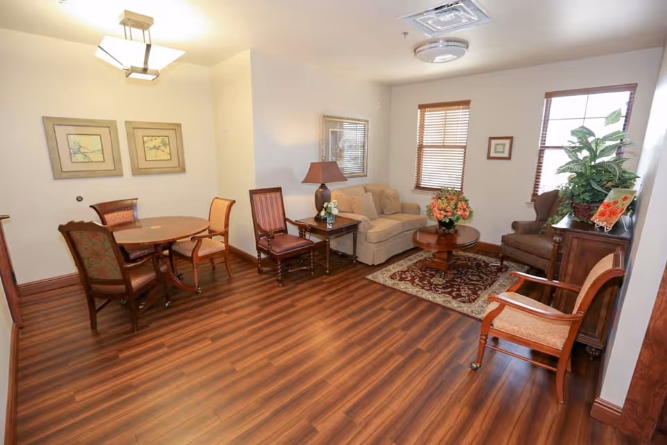 A bright, furnished common living area with wood floors, seating, a coffee table, dining table, and decorative plants and artwork.