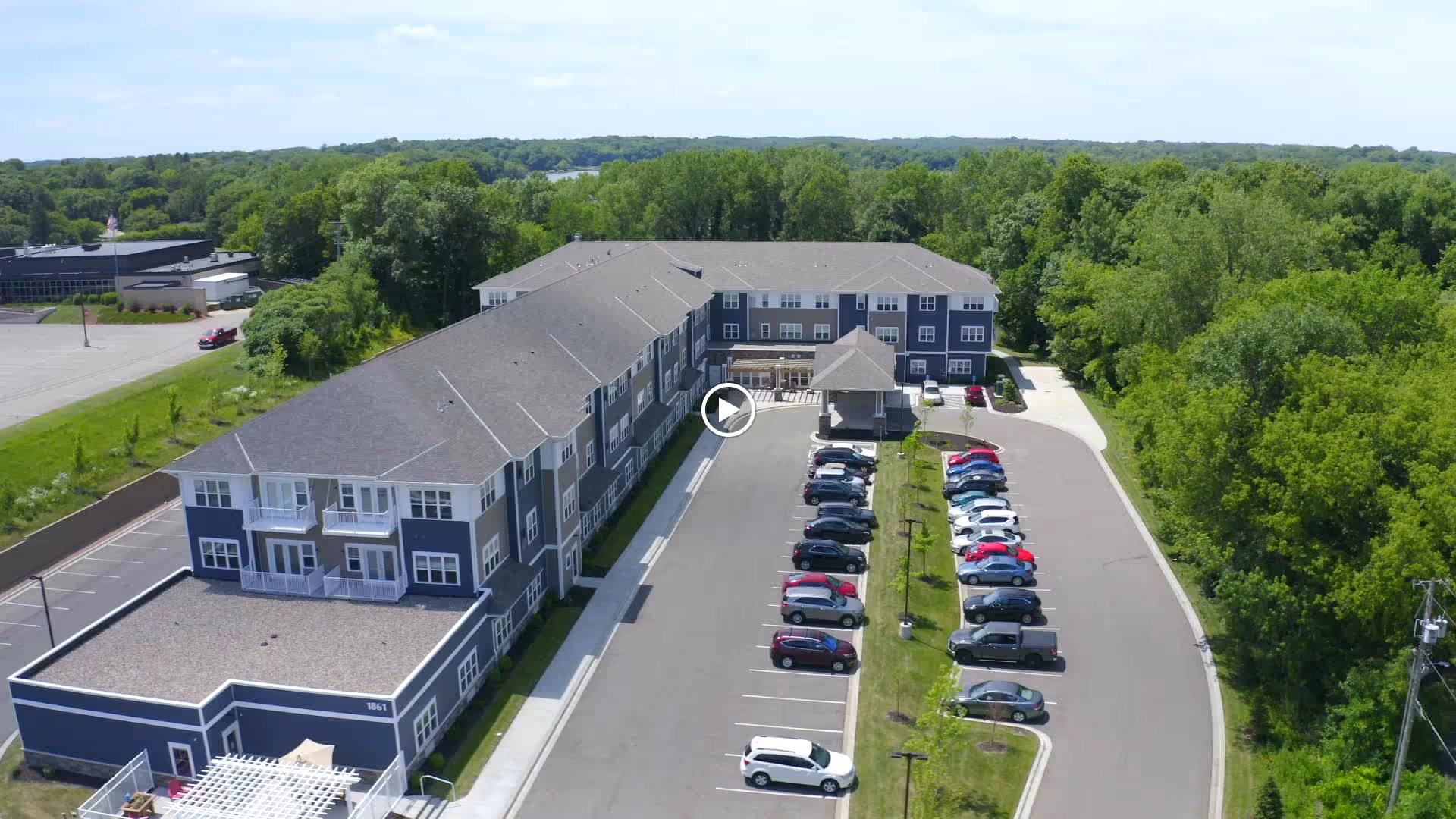 Aerial view of a three-story senior living building with a central covered entrance and a lined parking lot surrounded by trees.
