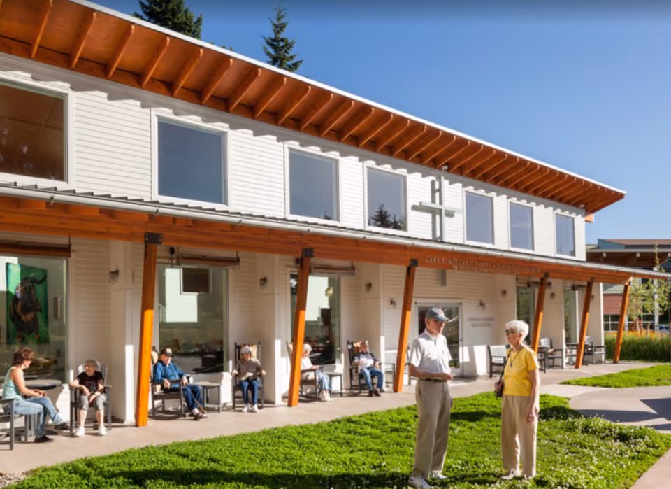 Outdoor patio area of a senior living facility with several elderly people sitting on rocking chairs under a wooden awning attached to a white two-story building. Two elderly individuals are standing and conversing on a grassy area in front of the building. The sky is clear and blue.