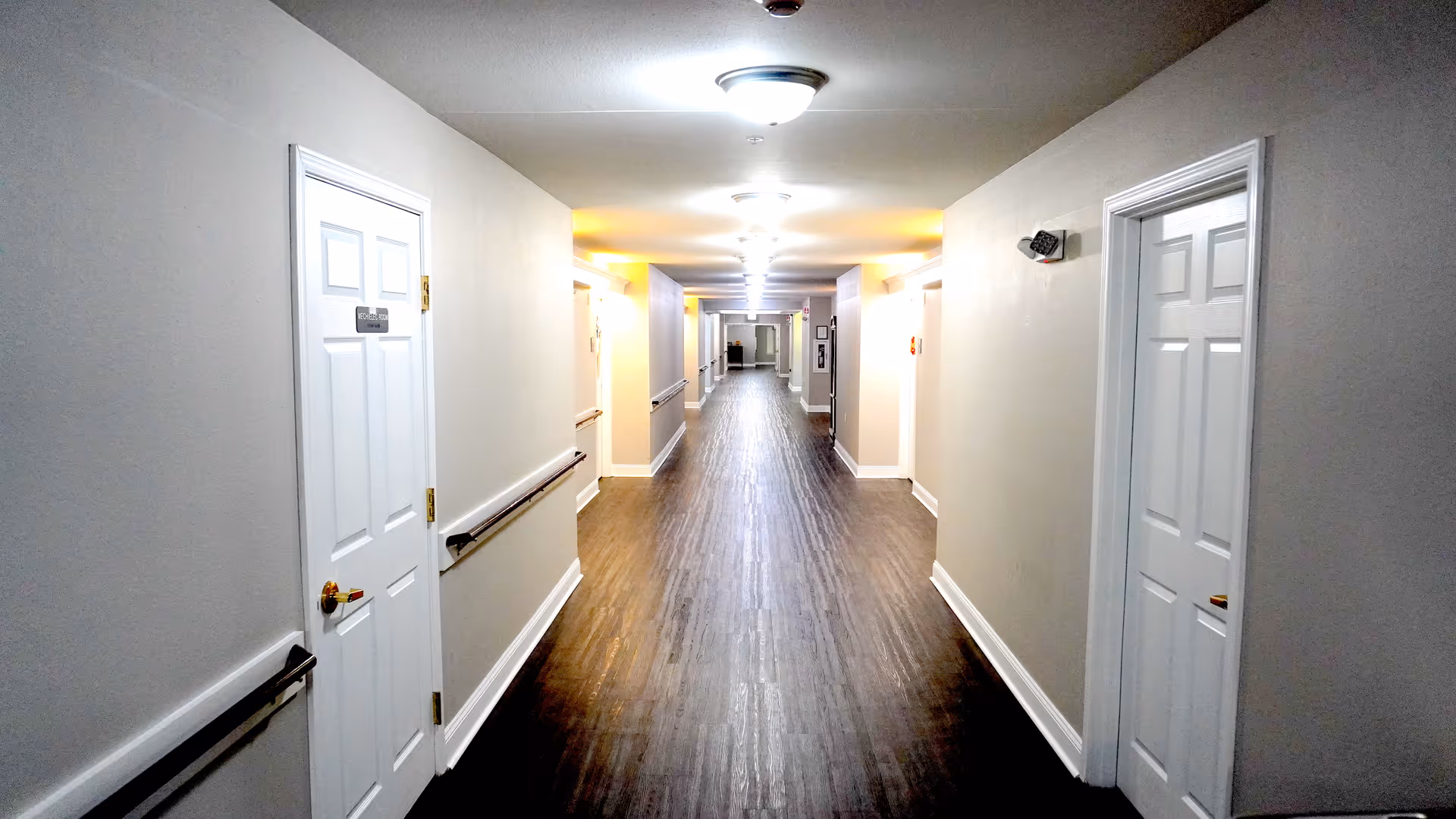 A long, well-lit hallway with wooden flooring and white walls. There are white doors on both sides of the hallway, each with a brass-colored door handle. Handrails run along both walls, and ceiling lights evenly illuminate the corridor.