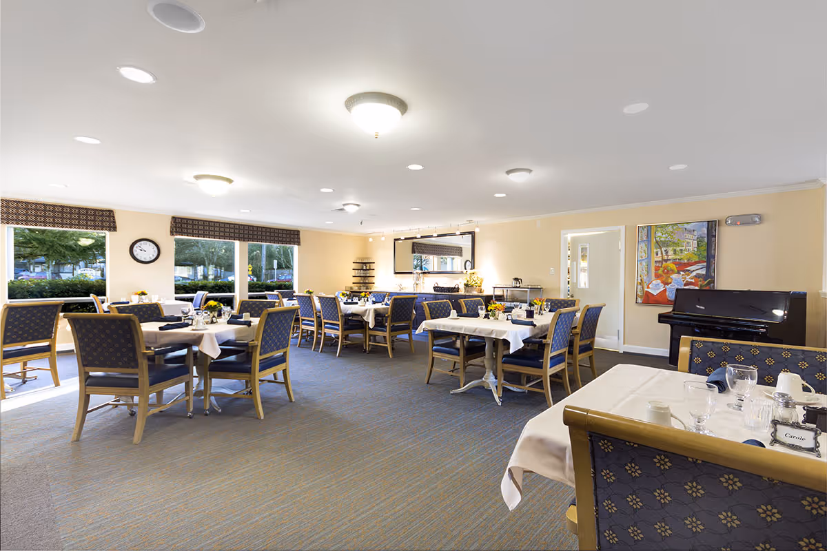 Bright senior living dining room with multiple set tables and chairs, large windows, and a piano in the corner.