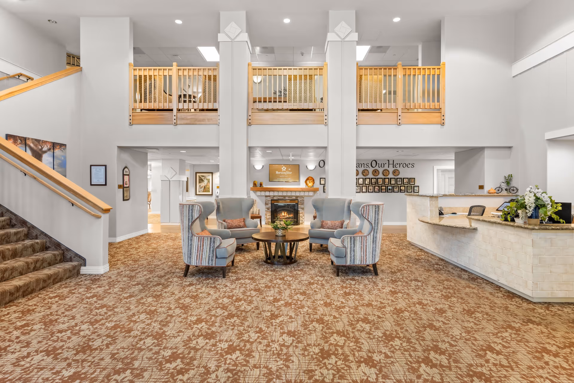Spacious senior living lobby with a seating area of armchairs around a table, a fireplace, reception desk, and a second-floor balcony.