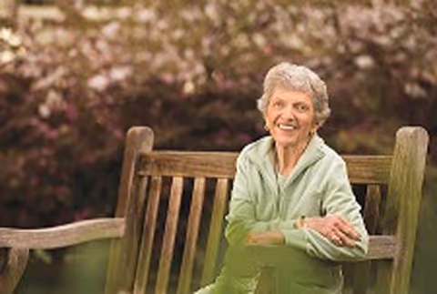 An elderly woman with short gray hair wearing a light green jacket sitting on a wooden bench outdoors with blurred trees and flowers in the background.